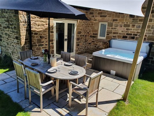a raised view of the garden patio in the sunshine, the table is set, the hot tub is open and bubbling in the background